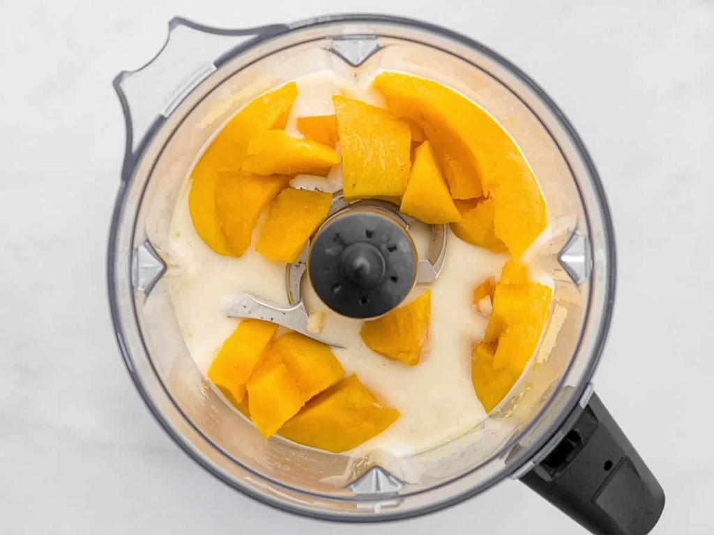 A high-speed blender filled with diced mangoes, coconut milk, lime